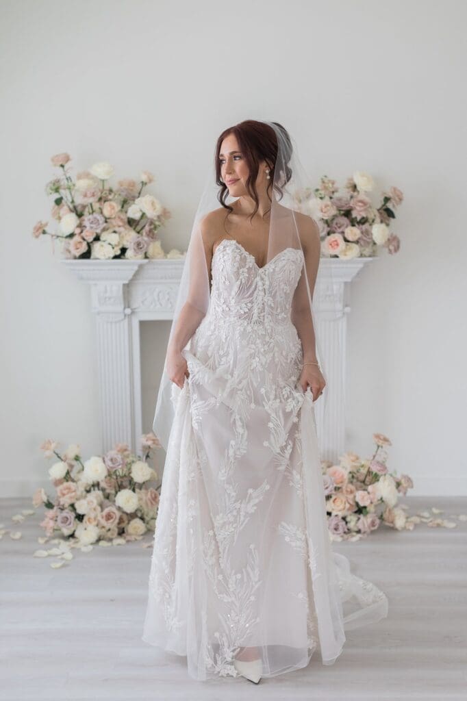 Bride looking out the window smiling, holding her dress. Her wedding dress is a sweetheart neckline, a line, and with a blush underlay and textured details over top