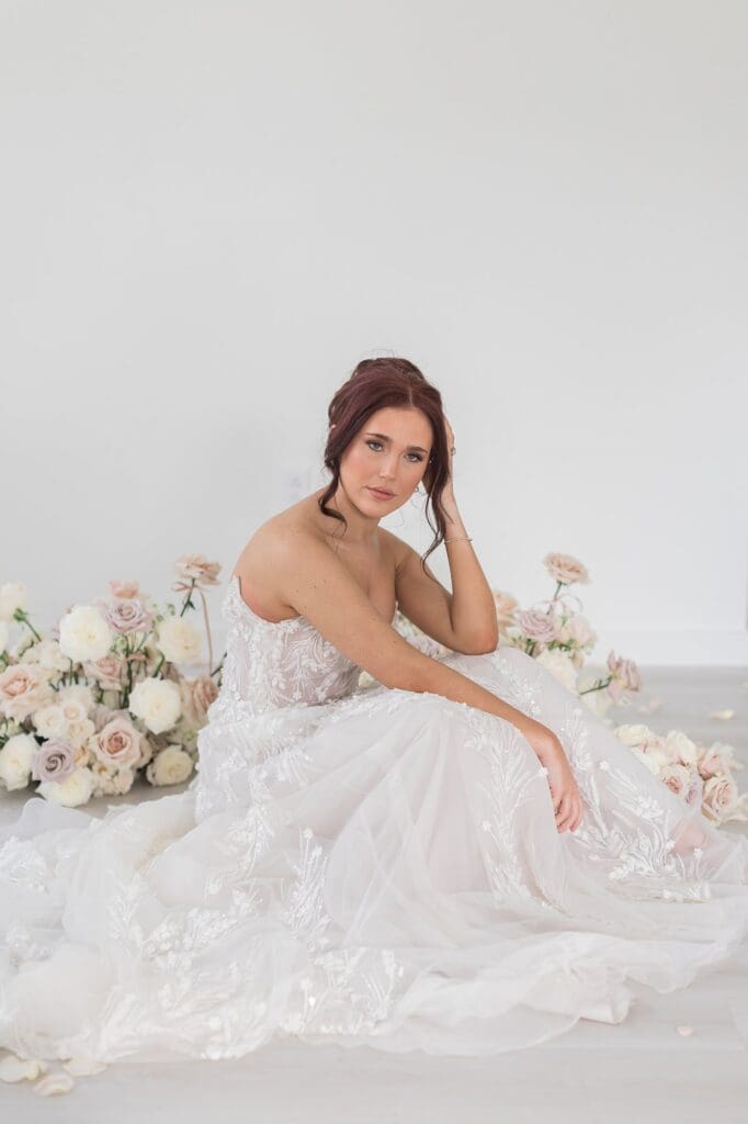 Editorial inspired shot of bride sitting among an arrangement of soft neutral florals. Her knees are slightly up to her chest with one arm draped over her legs and the other holding her head.