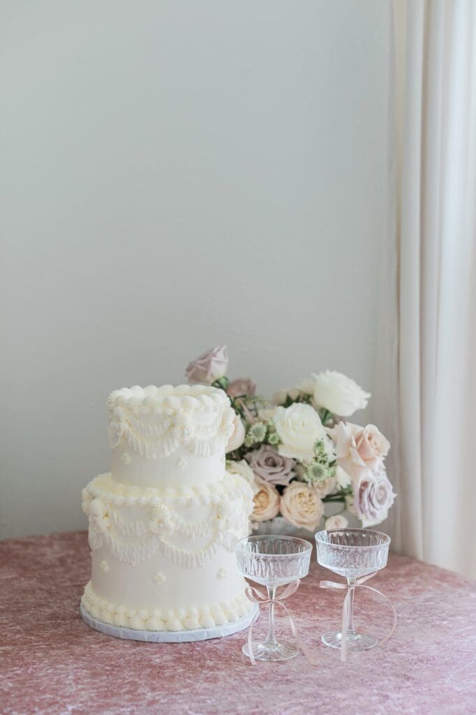 Two tiered white buttercream wedding cake with traditional design next to a floral arrangement and champagne glasses