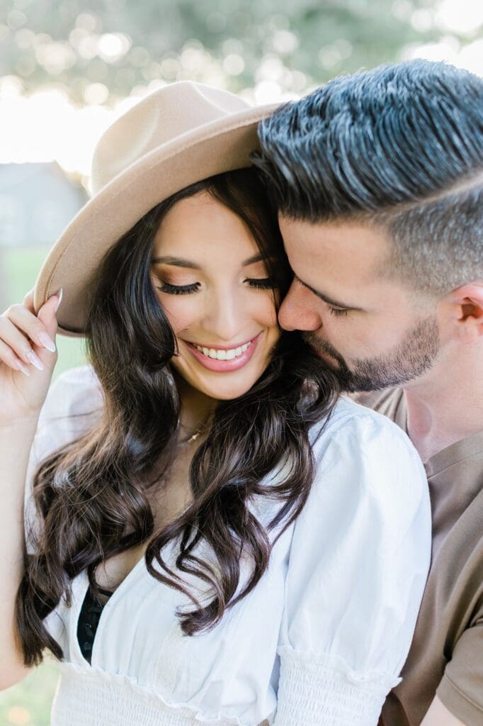 Bride holding her hat while the groom snuggles in for their engagement session at the orchard at caney creek