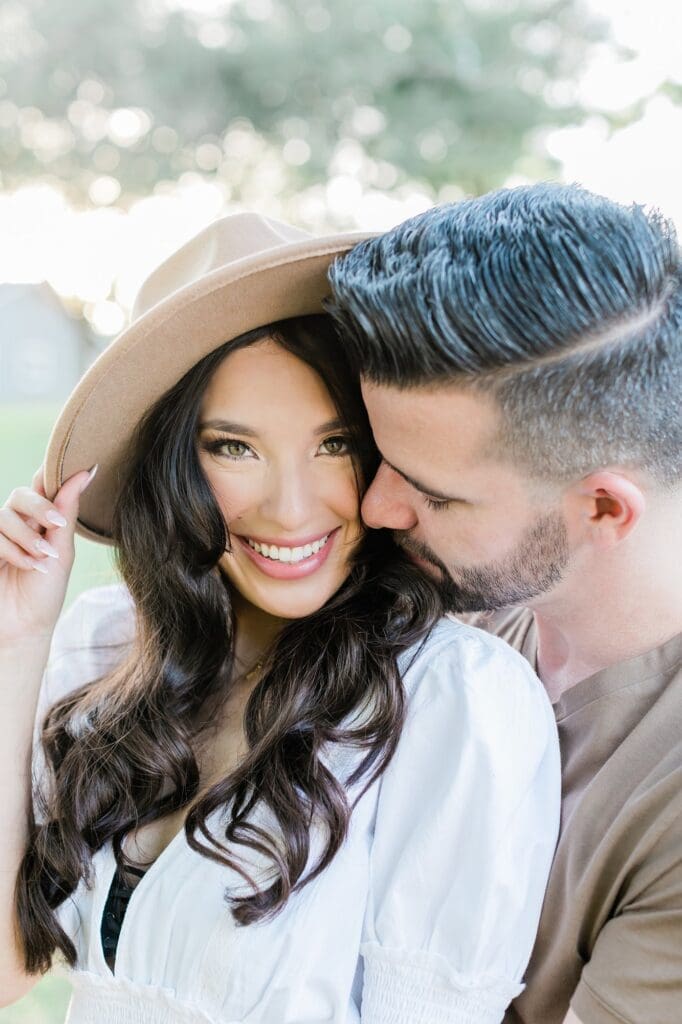 Bride holding her hat while the groom snuggles in for their engagement session at the orchard at caney creek