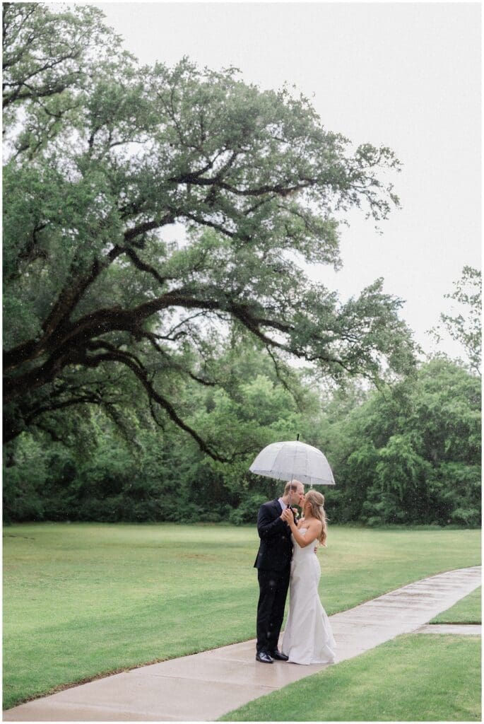 Bride and groom under the oak tree at Addison Woods. They are standing under a clear umbrella, sharing a kiss in the rain.