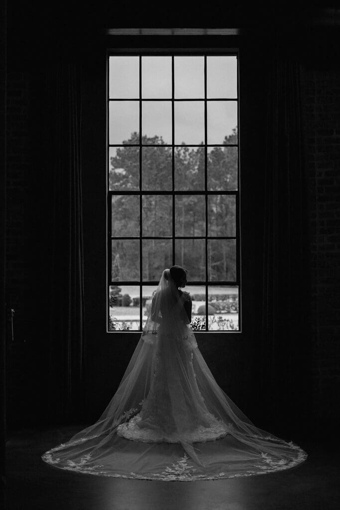 Black and white silhouette of bride standing in front of tall glass windows at Iron Manor 